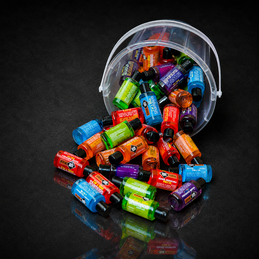 A clear plastic bucket tipped over on a dark reflective surface, spilling many small colorful bottles with dropper tops. The bottles contain bright orange, green, blue, purple, and red liquids and are arranged in a colorful pile.